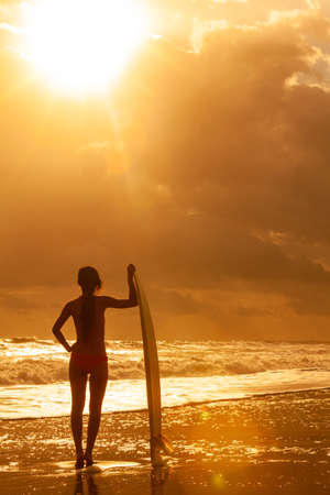 Rear view of beautiful sexy young woman surfer girl in pink bikini with white surfboard on a beach at sunset or sunriseの写真素材