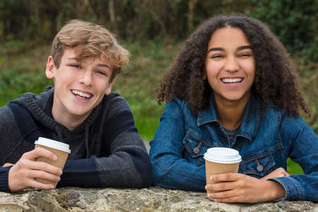 Mixed race group of two happy children teenagers, African American girl, caucasian boy leaning on a wall laughing and drinking takeout coffee togetherの写真素材