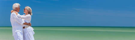 Panoramic banner image of a happy senior man and woman couple together embracing by sea on a deserted tropical beach with bright clear blue skyの写真素材