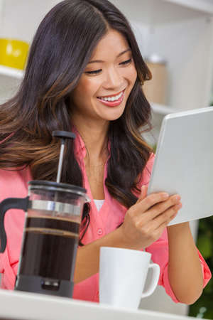 Beautiful young Asian Chinese woman or girl at home in her kitchen using a tablet computer drinking a cup or mug of coffee with a french press or cafetiereの写真素材