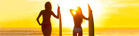 Panoramic web banner rear view silhouettes of beautiful sexy young women surfer girls in bikinis with surfboards on a beach at sunset or sunriseの写真素材
