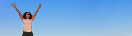 Panoramic web banner beautiful happy mixed race African American girl teenager female young woman celebrating arms raised smiling laughing with perfect teeth and sunglasses in front of a blue skyの写真素材