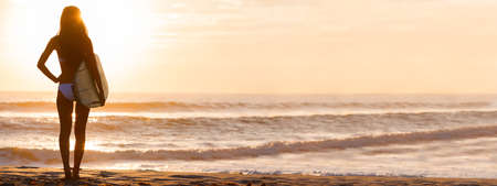 Panoramic rear view of beautiful sexy young woman surfer girl in bikini with white surfboard on a beach at sunset or sunriseの写真素材