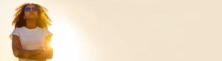 Panoramic web banner of beautiful mixed race African American female girl teenager young woman wearing aviator style sunglasses outdoors at sunset or sunriseの写真素材