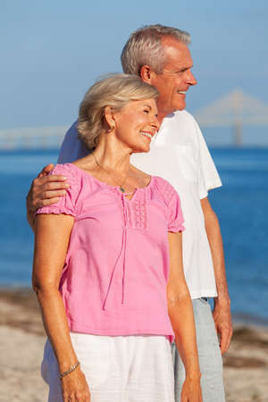 Happy senior man and woman couple standing embracing on vacation on a deserted white sand beach with bright clear blue sky and calm seaの写真素材