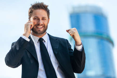 Successful business man, male executive businessman talking on his mobile cell phone celebrating in front of a high rise office block in a modern cityの写真素材