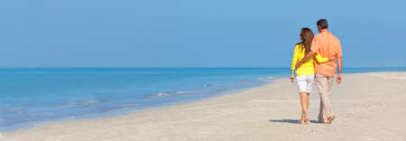 Web banner panorama rear view of man and woman romantic couple in colorful clothes walking on a deserted tropical beach with bright clear blue skyの写真素材