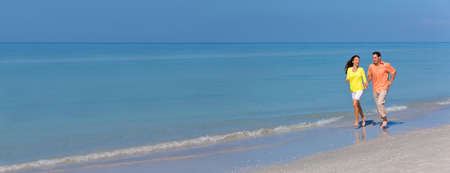 Panoramic web banner man and woman romantic couple running holding hands on a deserted tropical beach with bright clear blue skyの写真素材