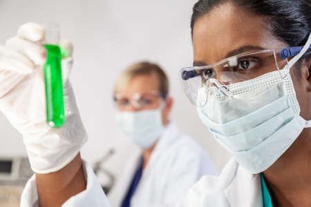 Asian medical or scientific researcher or doctor looking at a test tube of green solution in a lab or laboratory with her female colleague out of focus behind her.の写真素材