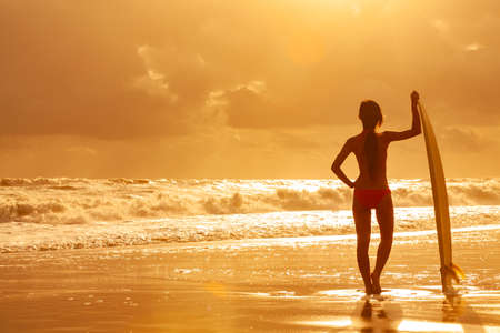 Rear view of beautiful sexy young woman surfer girl in bikini with white surfboard on a beach at sunset or sunriseの写真素材