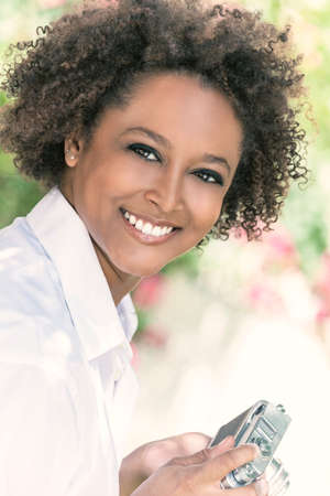 A beautiful mixed race African American girl or young woman looking happy taking pictures or photographs outside with a retro digital cameraの写真素材
