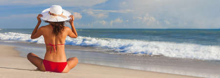 Pnoramic web banner rear view of beautiful young woman in red bikini and a white sun hat sitting on a deserted tropical beach with blue sky, healthy body care sun screen vacation conceptの写真素材