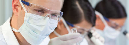 Panorama of male scientist, scientific researcher or doctor looking at a test tube of clear liquid in a medical research lab or laboratory with microscopes and his female colleagues behind him panoramic web imageの写真素材