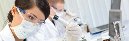 Panoramic web banner Chinese Asian female scientist, medical scientific researcher or doctor looking at a test tube of clear liquid in a research laboratory or lab with her colleague working behind her.の写真素材