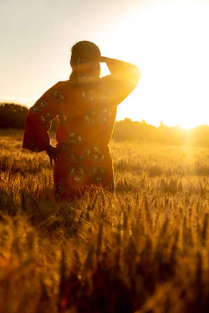 African woman in Africa wearing traditional clothes standing, looking, hand to eyes, in field of barley or wheat crops at sunset or sunriseの写真素材