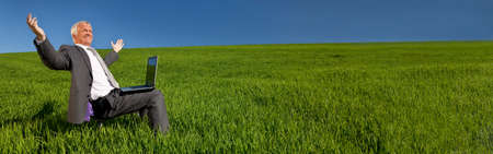 Panoramic web banner green environmental pension business concept of male businessman celebrating using a laptop computer in a green field with a blue sky. Panorama shot on location with copy space.の写真素材