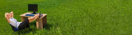 Panoramic web banner environmental business concept of a beautiful young woman sitting back relaxing at a desk with a computer in a green field with a bright blue sky with fluffy white clouds. Panorama on location with copyspace.の写真素材