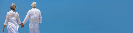 Panoramic web banner rear view of happy senior man and woman couple walking and holding hands with bright clear blue sky panoramaの写真素材