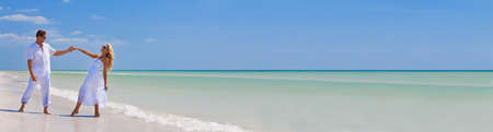 Panoramic web banner happy wealthy successful young man and woman couple dancing and holding hands on a deserted tropical beach with bright clear blue sky. Panorama header image.の写真素材