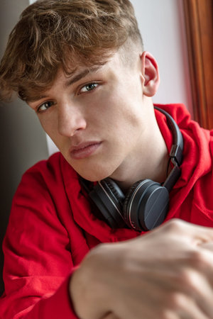 Serious moody male boy teenager young adult man indoors wearing a red hoodie and black wireless...