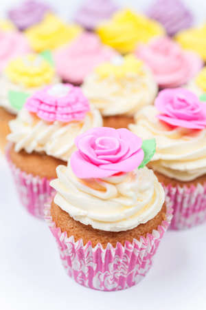Cup cakes with icing or frosting, pink, purple, yellow and cream with green leaves, rose and floral decorations photographed on a white backgroundの写真素材