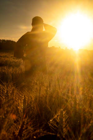African woman in traditional clothes standing, looking, hand to eyes, in field of barley or wheat crops at sunset or sunriseの写真素材
