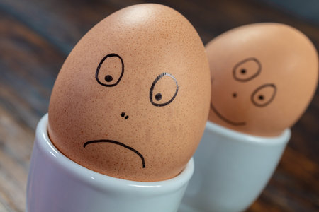 Emotions concept two boiled eggs in white egg cups with drawn happy and sad faces on a wooden tableの写真素材