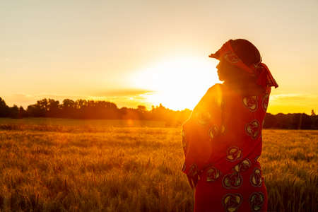 African woman in traditional clothes standing, looking, hand to eyes, in field of barley or wheat crops at sunset or sunriseの写真素材