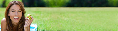 Panoramic beautiful woman in her thirties laying down outside on grass in a meadow or field holding eating an apple, smiling with perfect teeth. Dental or healthy eating concept web banner panoramaの写真素材