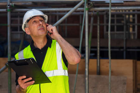 Worried or concerned male builder foreman, worker, contractor or architect on construction site holding black clipboard and talking on his cell phoneの写真素材