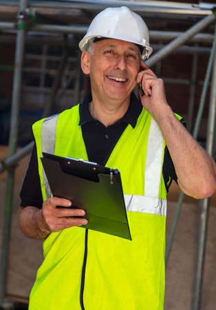 Male builder foreman, worker, contractor or architect on construction site holding black clipboard and talking on his cell phoneの写真素材