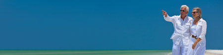 Panoramic web banner happy senior man and woman couple walking together looking out to sea on a deserted tropical beach with bright clear blue sky, the man is pointing to the horizon panorama imageの写真素材