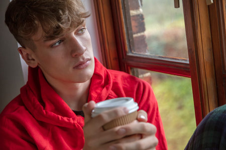 Sad depressed thoughtful teen boy male teenager young man looking out of a window drinking coffee or teaの写真素材