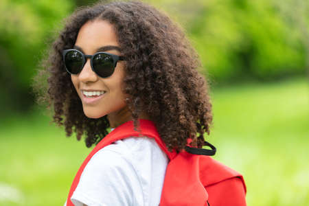 Outdoor portrait of beautiful happy mixed race biracial African American girl teenager female young woman smiling laughing with perfect teeth hiking with red rucksack and wearing sunglasses in green countrysideの写真素材