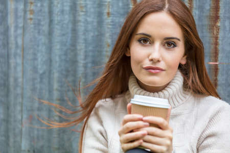 Outdoor portrait of beautiful girl or young woman with red hair wearing a white jumper drinking takeout coffeeの写真素材