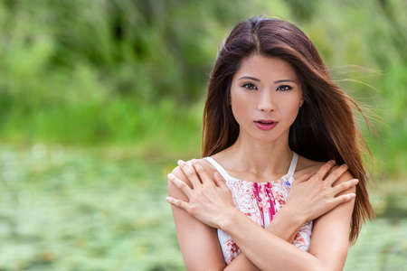 Outdoor portrait of a beautiful young Chinese Asian young woman or girl with perfect make up arms crossed over her chest in front of a natural green backgroundの写真素材