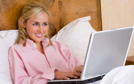 Beautiful young woman female teenager laying in bed wearing a pink short and using her laptop computer on the internet or on line for social mediaの写真素材