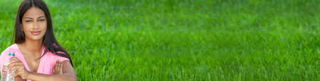 Panoramic outdoor portrait of a beautiful happy Indian Asian young woman or girl outside in summer sunshine drinking from a bottle of water. Panorama web banner.の写真素材