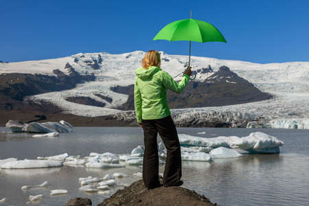 Climate change concept woman female hiker holding a green umbrella standing on a rock looking out on a melting glacier and icebergs in Icelandの写真素材