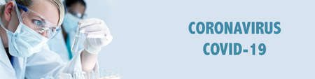 Panorama female woman medical or scientific researcher or doctor wearing a face mask looking at a test tube in a laboratory with her Asian colleague out of focus behind her. Panoramic web banner with Coronavirus text.の写真素材