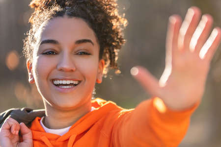 Outdoor portrait of beautiful happy mixed race African American girl teenager female young woman laughing, waving and smiling with perfect teeth in golden evening sunshineの写真素材