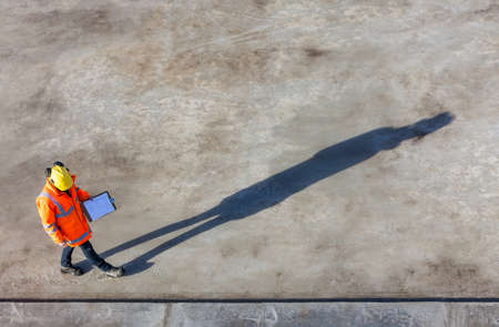 Overhead aerial view of anonymous construction worker or builder wearing a hi vis jacket, a hardhat and carrying a clipboard on a building site with their shadow stretching out in front of themの写真素材