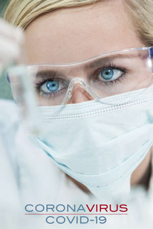 Female woman medical scientific researcher or doctor wearing a face mask looking at a sample test tube in a Coronavirus COVID-19 lab or laboratory. Photograph with text.の写真素材