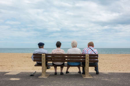 Rear view of four anonymous senior people, old couples, elderly men and women sitting on a bench at a beachの写真素材