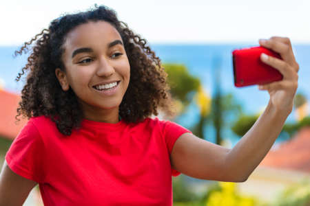 Beautiful biracial mixed race African  teenager teen girl young woman smiling with perfect teeth taking selfie on cell phone outside by the sea in summerの写真素材
