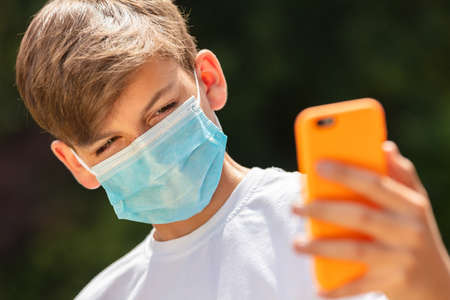 Boy teenager teen male child wearing a face mask outside during the Coronavirus COVID-19 virus pandemic while using his mobile cell phone for a selfie for social mediaの写真素材