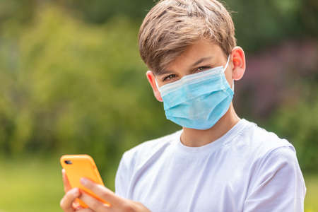 Boy teenager teen male child wearing a face mask outside during the Coronavirus COVID-19 virus pandemic while using his mobile cell phone for social media or text messagingの写真素材
