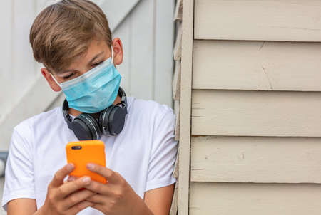 Boy teenager teen male child wearing a face mask outside during the Coronavirus COVID-19 virus pandemic while using his mobile cell phone and wireless headphonesの写真素材