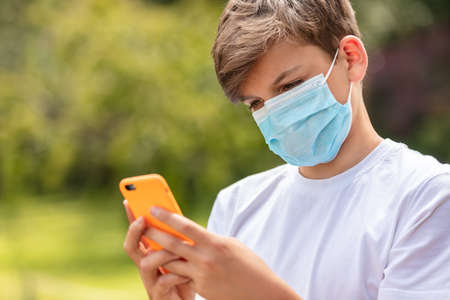 Boy teenager teen male child wearing a face mask outside during the Coronavirus COVID-19 virus pandemic while using his mobile cell phone for social media or text messagingの写真素材