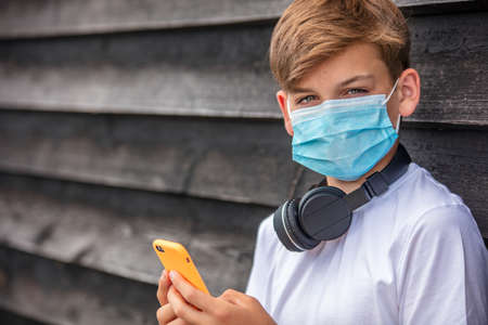 Boy teenager teen male child wearing a face mask outside during the Coronavirus COVID-19 virus pandemic while using his mobile cell phone and bluetooth wireless headphonesの写真素材